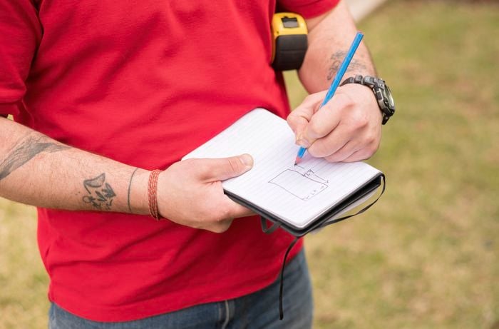  A person in a red T-shirt is sketching a labeled diagram of length and width in a notebook while holding a pencil and measuring tape. 