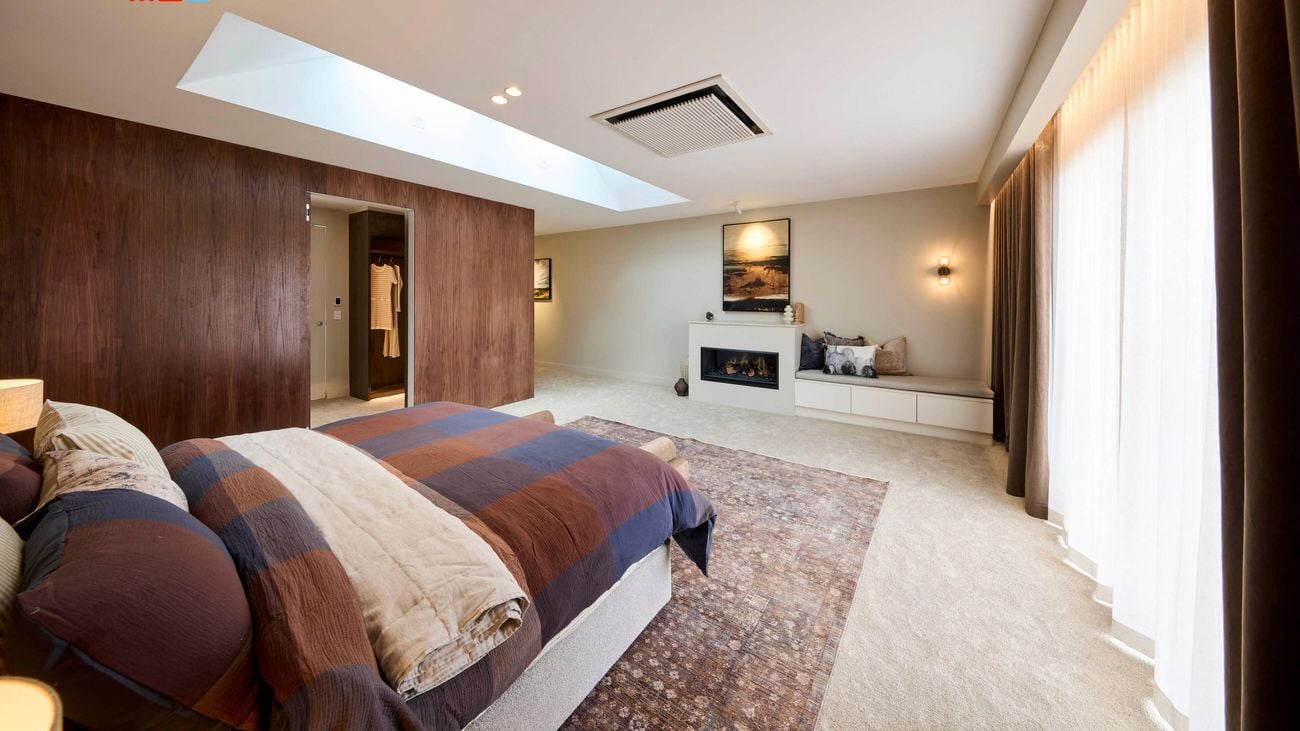 A spacious, modern bedroom with a large bed dressed in a plaid blanket and pillows. The room includes a skylight, a wooden accent wall, a built-in fireplace with a seating area, and large windows with curtains that allow natural light to enter.