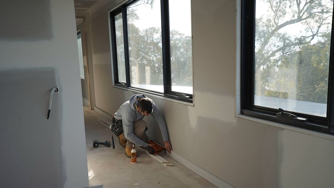 A tradesperson uses a Paslode nailer and flooring adhesive to secure floor trims in a building hallway before flooring installation.