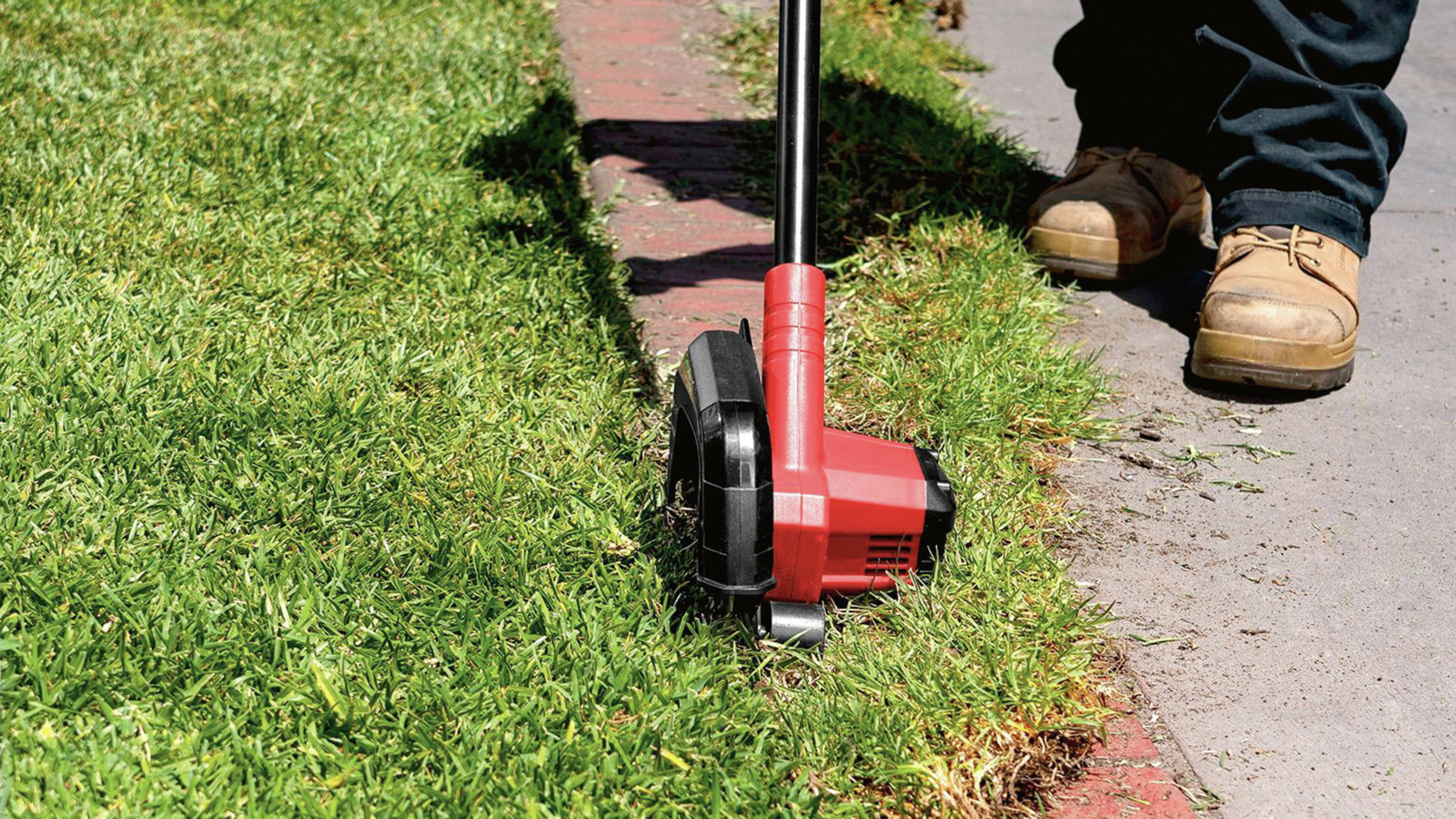 A person wearing tan work boots and dark pants uses a red and black lawn edger to trim the grass along the edge of a concrete footpath and brick border.