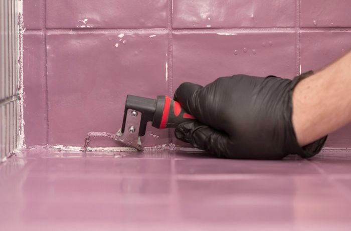 A person wearing black gloves is using a silicone removal tool to scrape silicone from the edges of tiles.  ]    