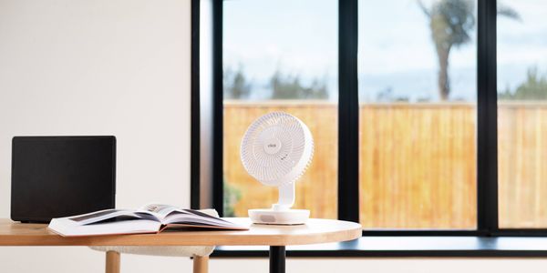 Compact white desk fan positioned on a home office desk beside an open book and laptop in front of large windows.