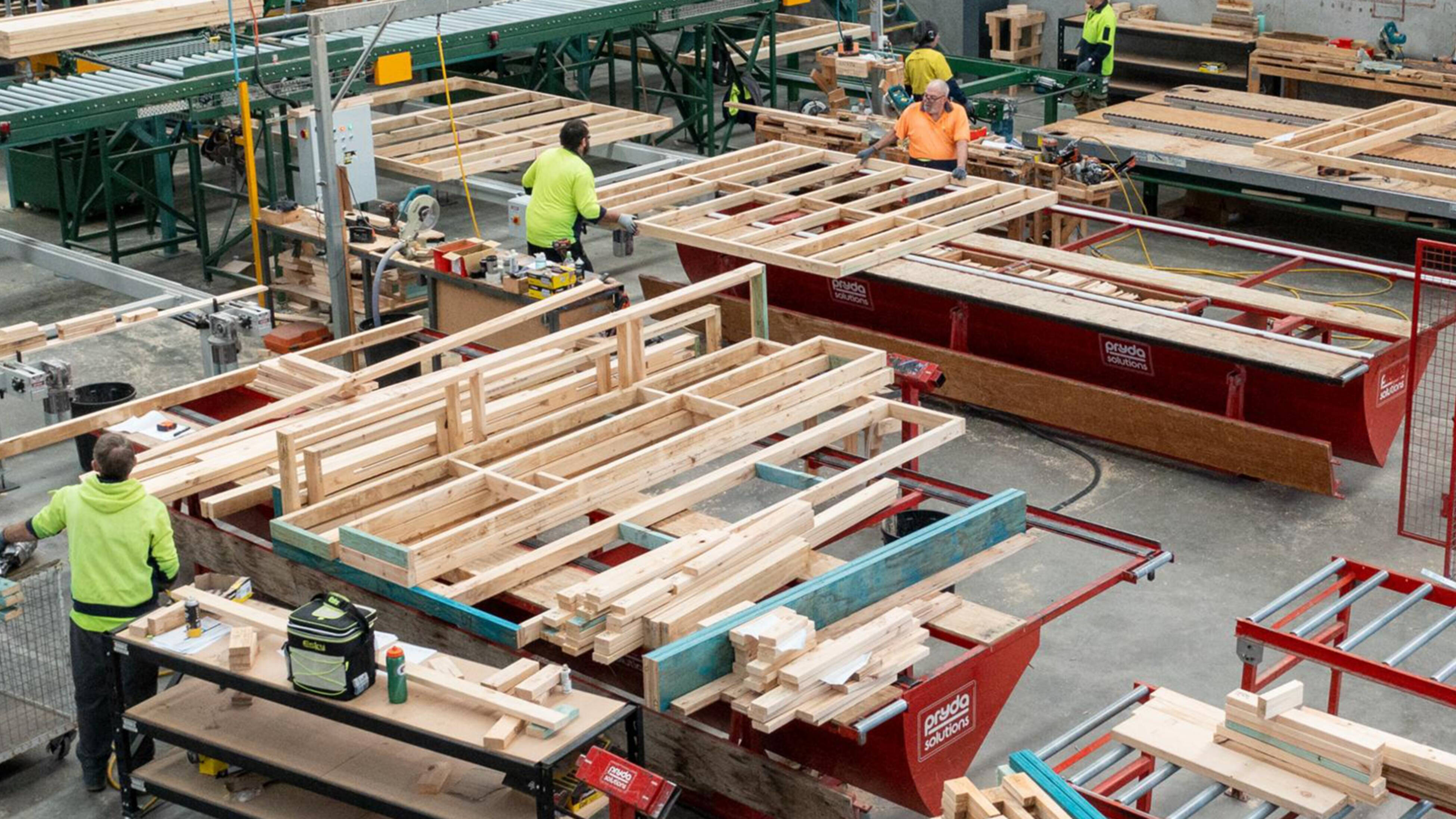 Workers assembling prefabricated timber frame and truss on workstations in a factory with tools and materials visible.