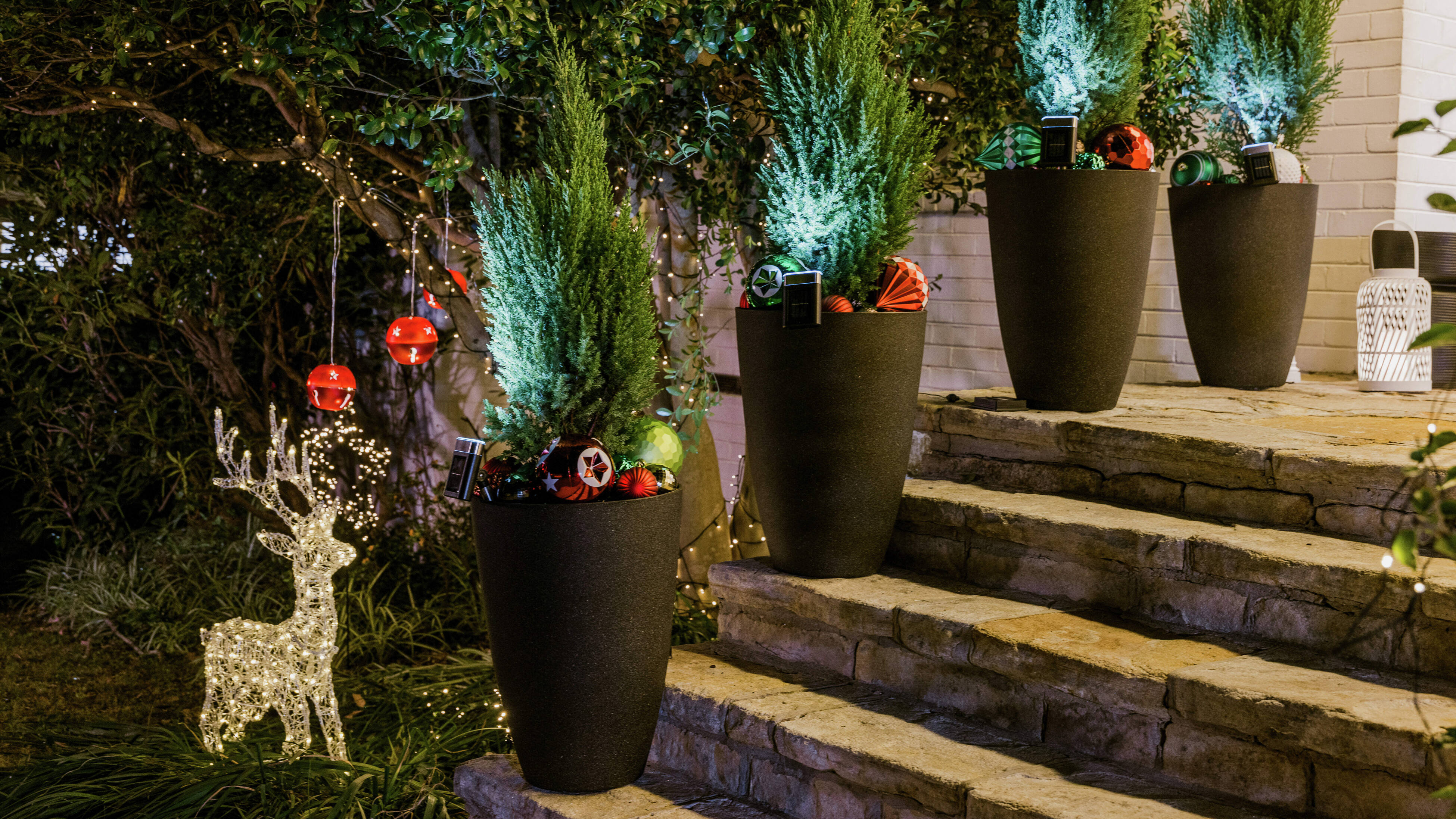 Four tall planters with greenery and ornaments on steps, surrounded by fairy lights and a glowing reindeer decoration.