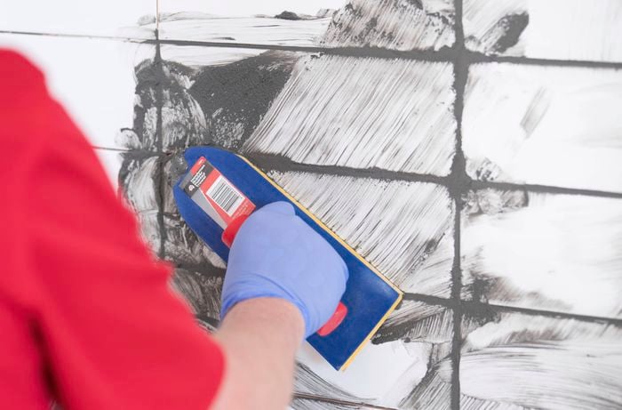 A person wearing orange gloves is laying the white tiles on the wall using spacers to keep the gaps even.  