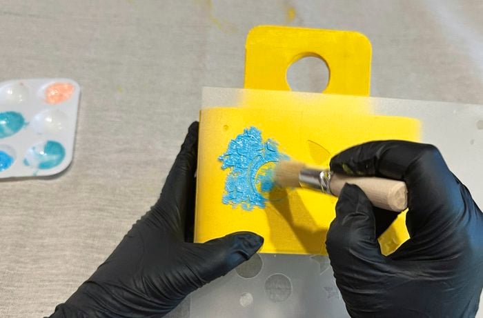 Hands wearing black gloves using a stencil and a brush to apply blue paint onto the yellow art caddy, creating a moon and star design.