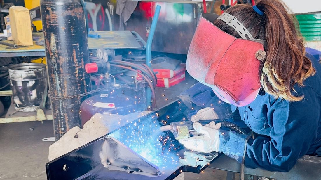 A person wearing protective welding gear, including a large face shield and gloves, is welding a black metal component on a workbench. Bright sparks and blue light from the welding torch illuminate the workspace. The surrounding area contains gas cylinders, tools, and industrial equipment, indicating a workshop environment.