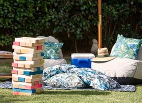Giant completed tumbling tower game stacked up in an outdoor setting with large cushions, table, hat and esky under an umbrella