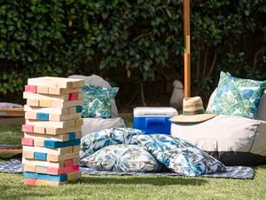 Giant completed tumbling tower game stacked up in an outdoor setting with large cushions, table, hat and esky under an umbrella