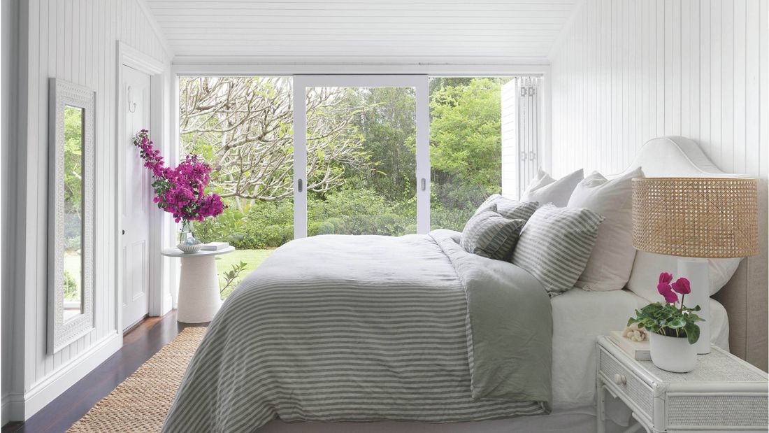A bright bedroom with a bed a decorative full-length mirror on the wall and a large window looking out onto a lush green garden.