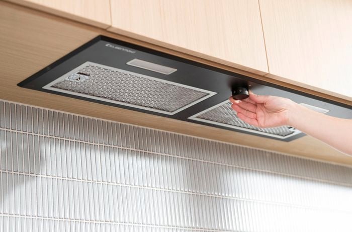 A person adjusts the control knob on a modern stainless steel rangehood mounted under kitchen cabinets.  
