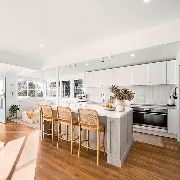 Bright open-plan kitchen and living area with a large island bench, white cabinetry, timber flooring, woven bar stools and sunlight streaming through open doors and windows.