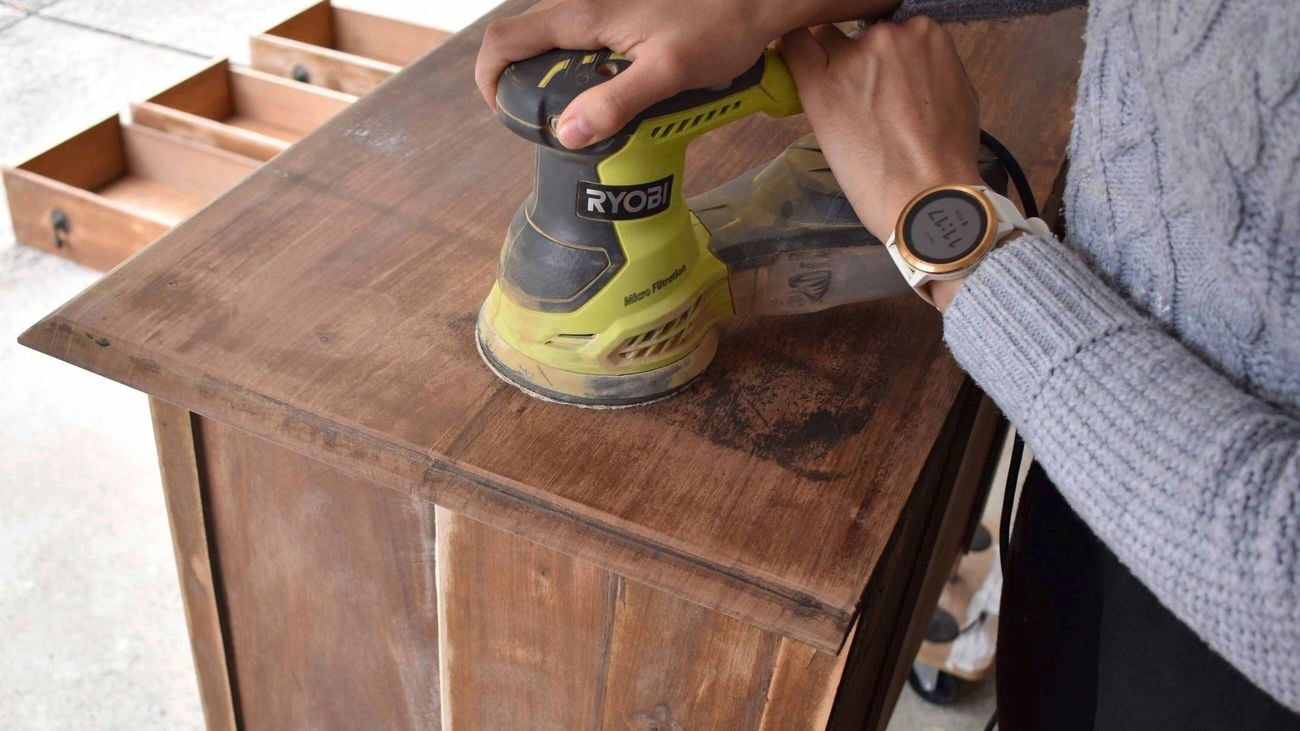 A person using a sander to sand a wooden dresser.