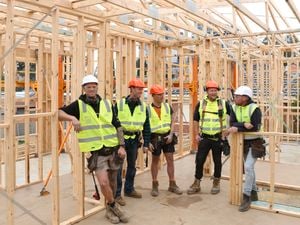 Five carpenters, two in orange hard hats and three in white hard hats, stand amongst frame and truss work onsite