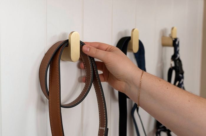 A person is hanging a brown dog leash on a gold wall hook, with other leashes already hanging nearby.