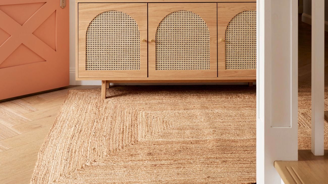 Brown rug running from front door entrance with staircase in foreground and wooden cabinet in background.