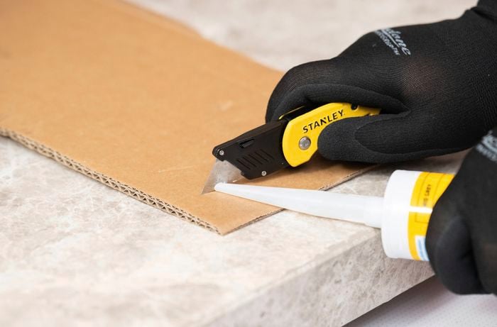 Black-gloved hand uses a utility knife to trim the nozzle of a silicone cartridge while the cartridge rests on a stone-look benchtop beside a piece of cardboard.