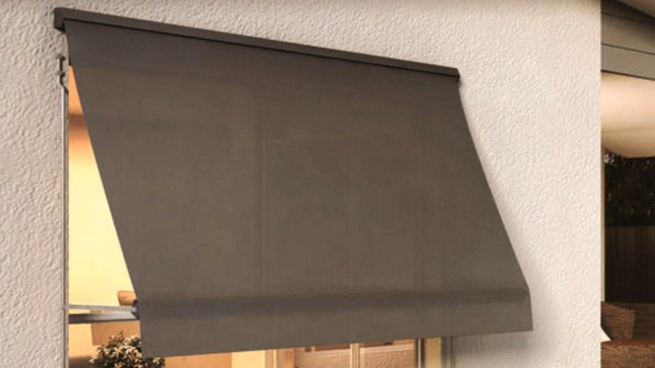 A black Windoware awning installed outside over a window above a garden bed, shading a lit living room.