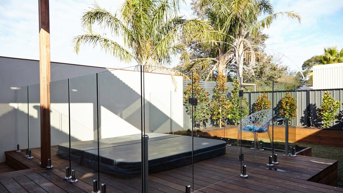 A small backyard complete with spa, deck with glass pool fence, and trees lining the back fence