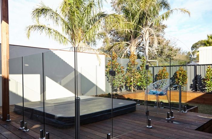 A small backyard complete with spa, deck with glass pool fence, and trees lining the back fence