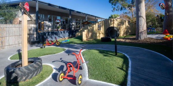 The outdoor playground of a kindergarten, with tricycle, concrete track, tanbark area and big tree, in front of a large building.