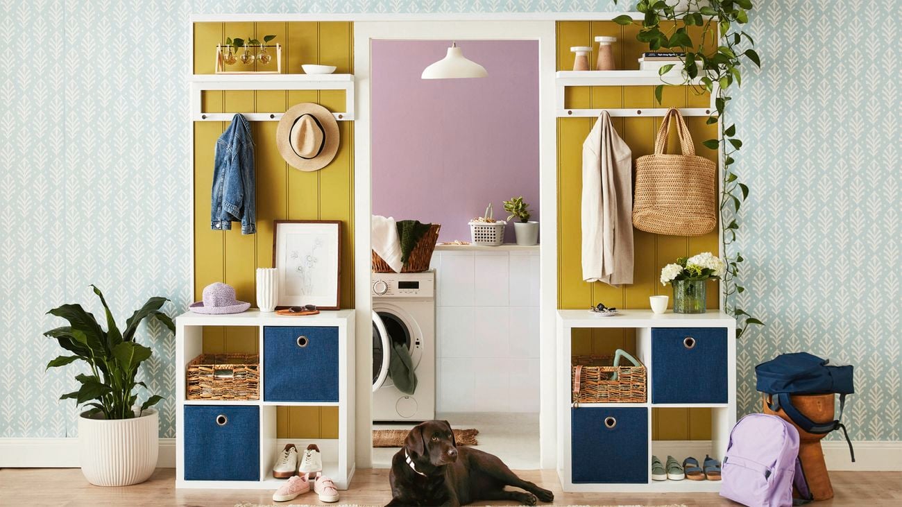 An entryway with a yellow wall and white cube shelving holding a straw hat, denim jacket, and woven baskets. A black Labrador dog lies on the floor, surrounded by shoes and a purple backpack. In the background is a laundry room with a washing machine and purple wall.