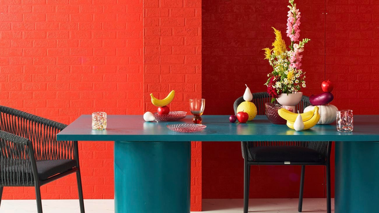 Dining room setting with teal dining table, hanging lights and a red and pink brick wall as a backdrop.