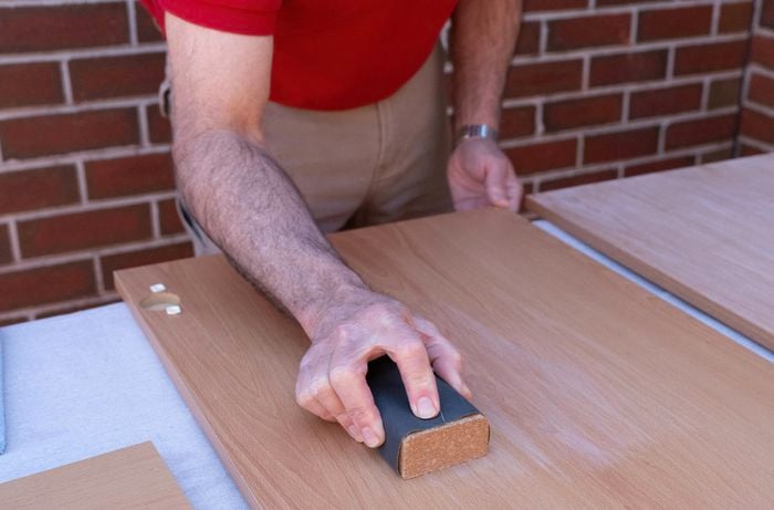 A person is sanding cabinet doors, drawers, and surfaces with a sanding block to prepare the laminate for primer and paint