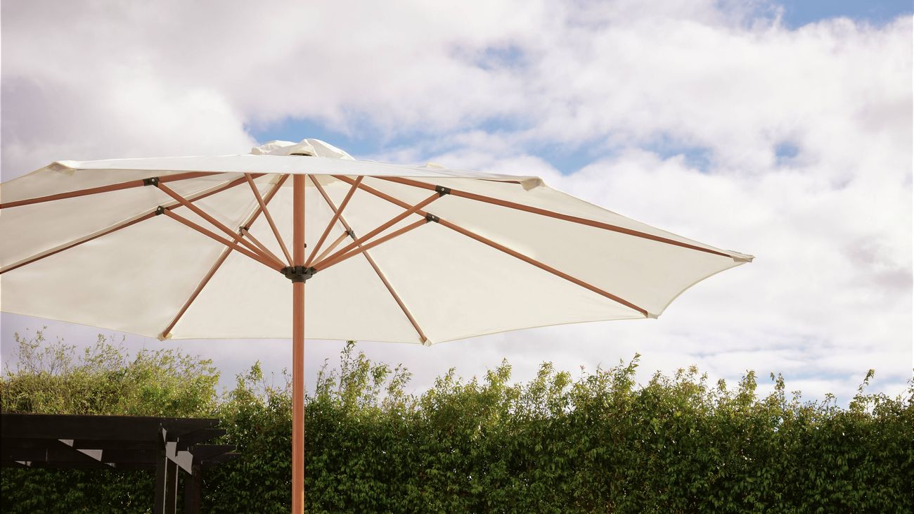 An L-shaped white outdoor lounge set with wooden frame and coffee table in a garden, with a shade umbrella propped behind.