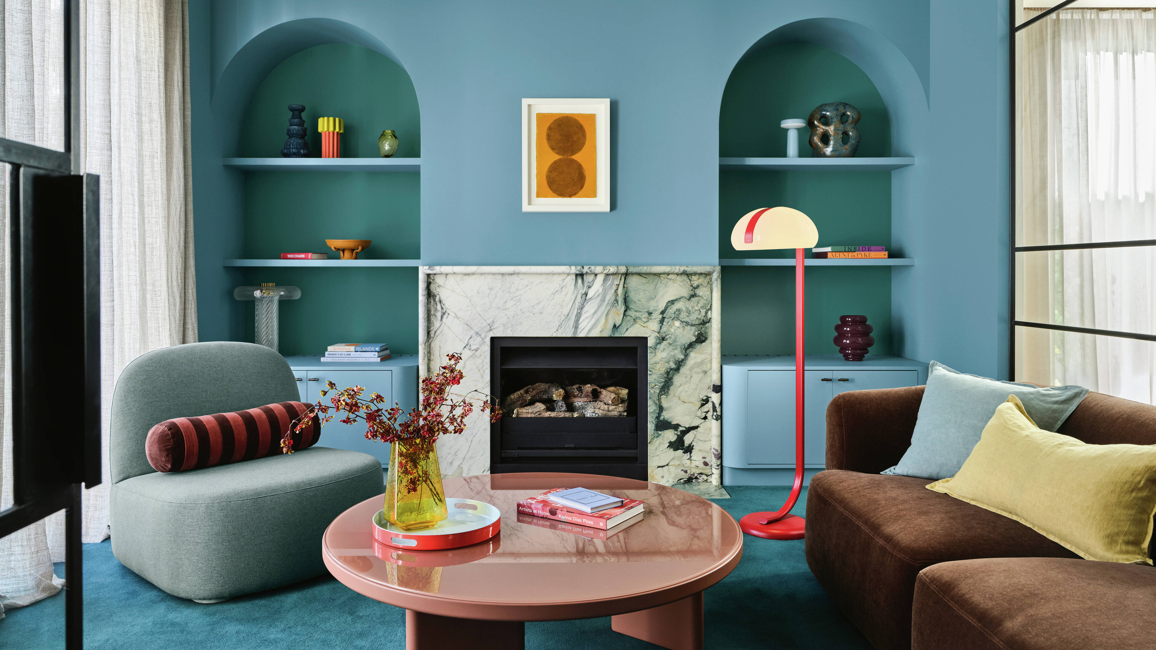 Modern living room with blue walls and arched built‑in shelves, a marble‑veined fireplace topped with abstract artwork, blue carpet, a round pink gloss coffee table with a vase and books, a grey curved lounge chair with a striped bolster cushion, a brown sofa with a mustard cushion, and a red floor lamp.