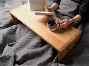 A pair of hands resting on a timber breakfast table holding a book and cup of tea from bed