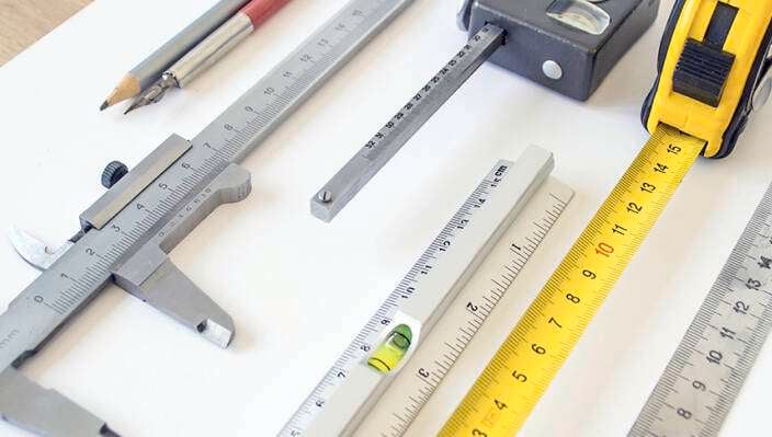 A collection of measuring tools including a caliper, metal rulers, a spirit level, a pencil, a square, and a yellow tape measure arranged on a white surface.