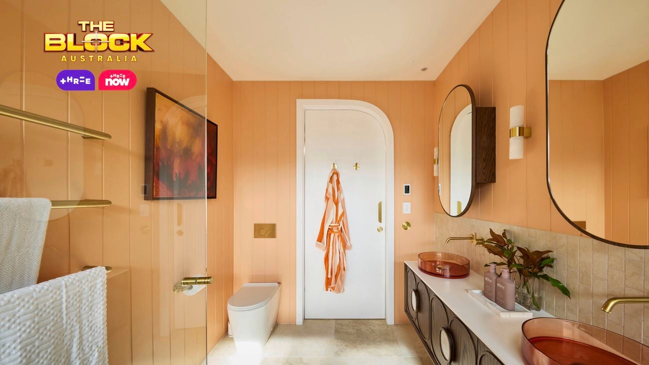 Modern bathroom with peach walls, double copper sinks, round mirrors, gold fixtures, white toilet, and orange robe on door.