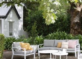  An outdoor seating area with grey and white furniture, cushions and a round table, surrounded by greenery and a large tree near a white house