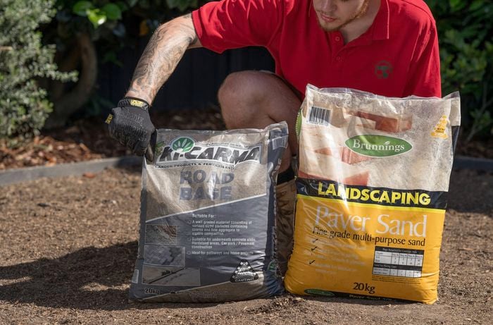 A Bunnings Team Member is calculating the correct depth to dig, holding a bag of road base while next to a bag of landscaping paver sand.