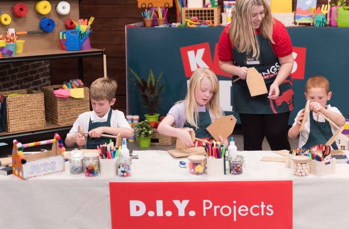 Three kids are doing DIY projects with a Bunnings team member. On the table, there is a decorated tool caddy and many arts and crafts supplies.