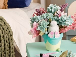 A painted plant pot decorated with an Easter theme is filled with artificial flowers and a small bunny figure, sitting on a craft table in a bedroom setting.