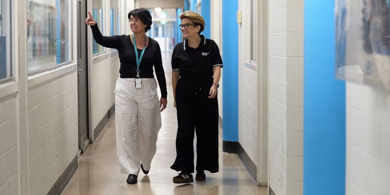 Person in a Bunnings Trade polo walks through an educational facility hallway with another person, pointing towards a classroom window.
