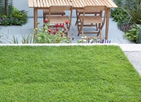 A patch of neat and lush green grass in the foreground. Behind the grass is a sunken grey stone patio with a wooden table and chairs and colourful flowers edging the patio borders