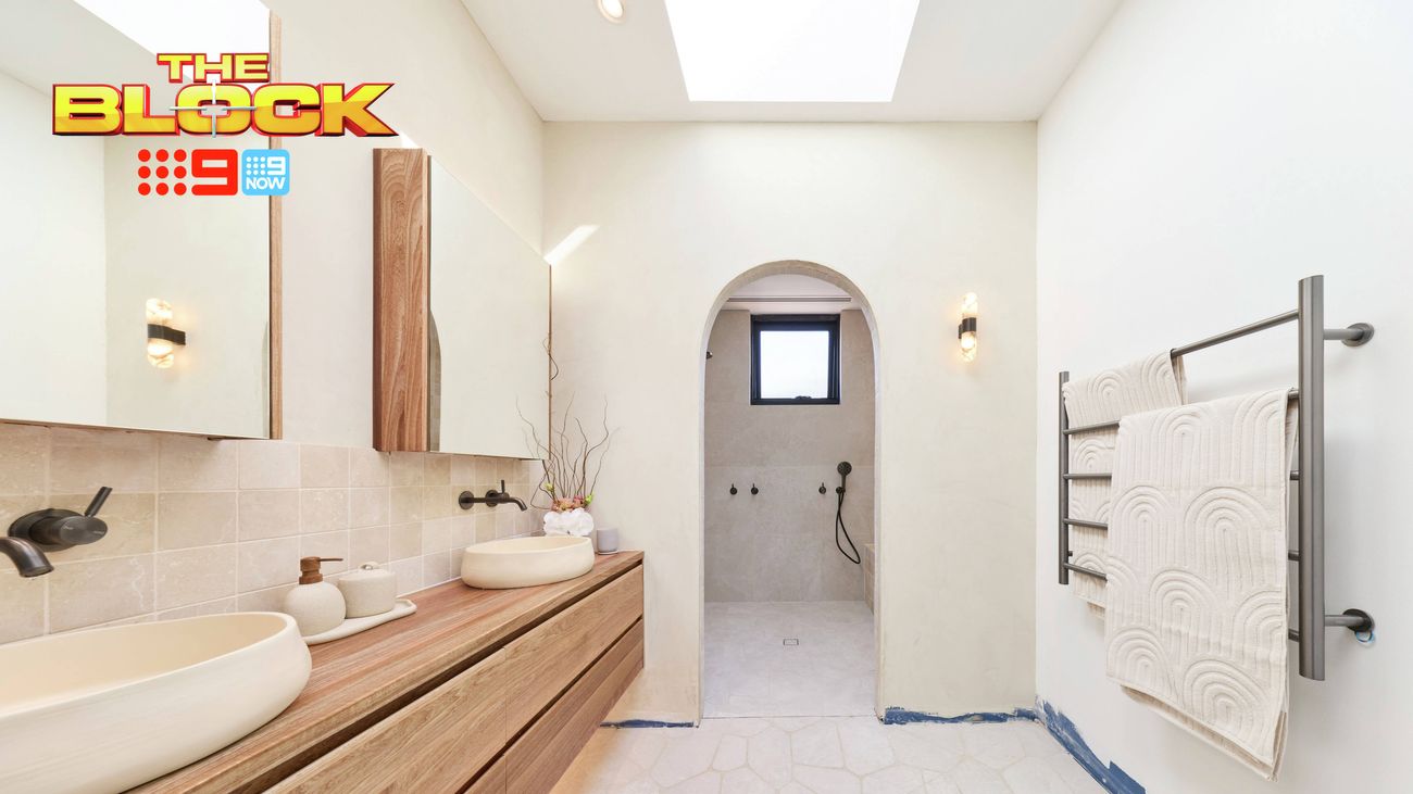 Modern bathroom with a long timber vanity, white vessel basins, large mirrors, black tapware, heated towel rail and an arched doorway leading to the shower. “The Block” logo appears in the upper-left corner.