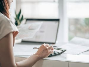 A lady typing on a calculator on a desk with a laptop and some pieces of paper on it