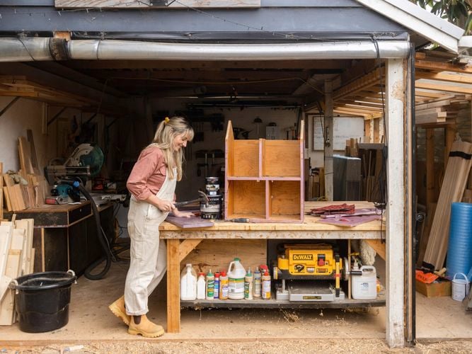 A lady working in and outdoor shed, using a hammer to fix broken timber doll house