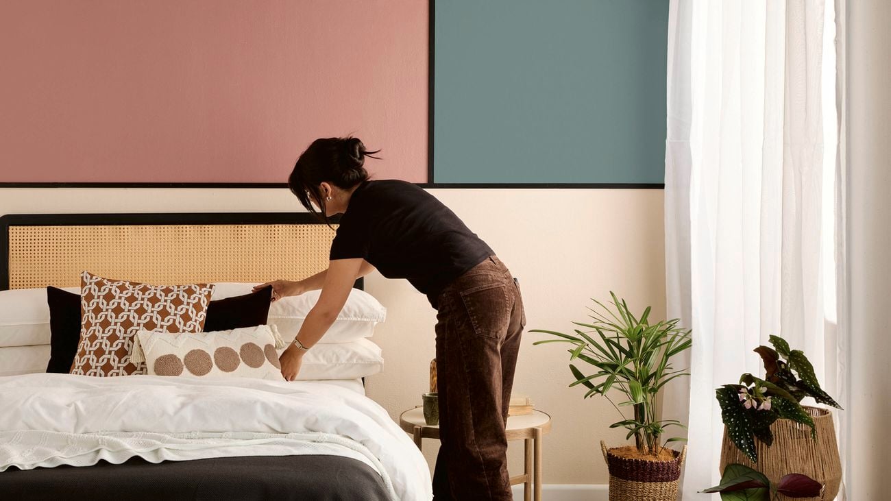 A woman adjusting pillows on a bed with white bedding, patterned cushions, and a black throw, against a cane headboard. Behind is a pink and green colour‑blocked wall with black trim.