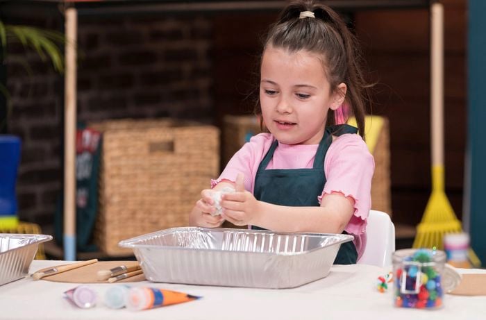 A young girl in a pink shirt and apron is preparing the clay with her hands, with a BBQ tray underneath to protect the table.