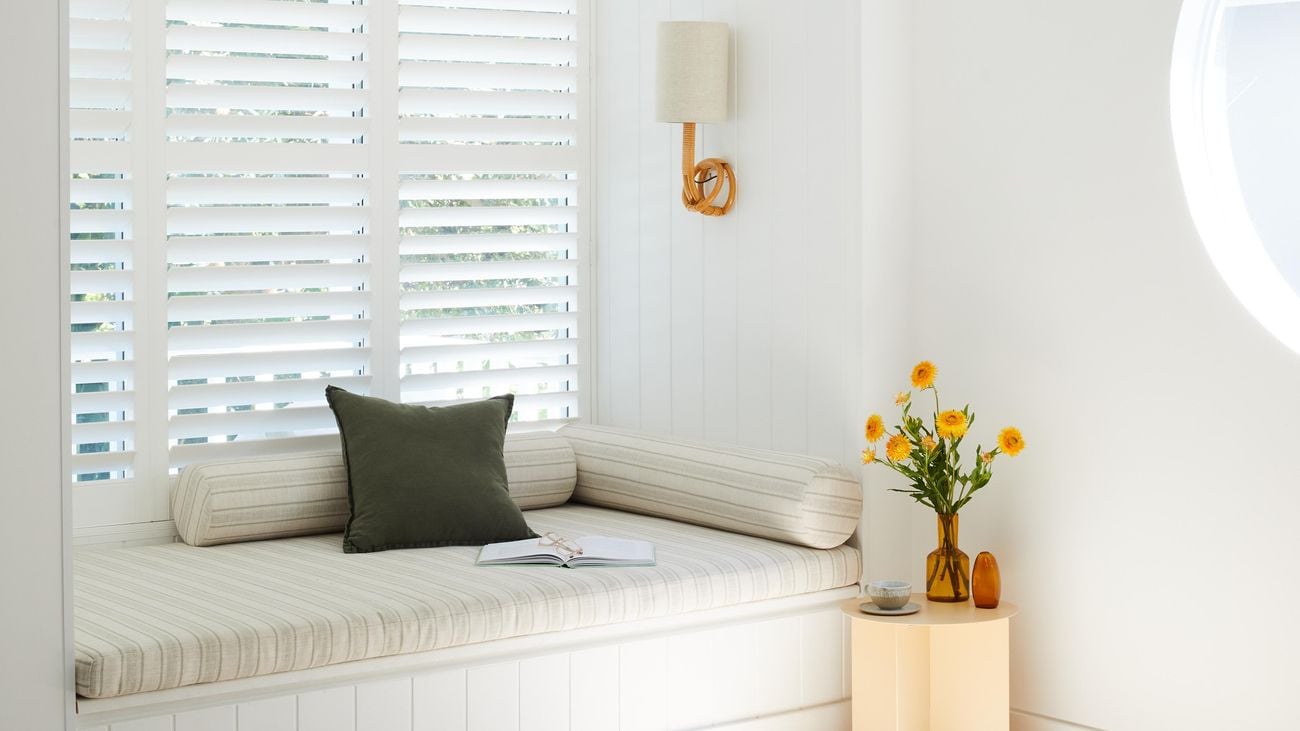A vase of yellow flowers sit on top of a small, circular table in the corner of a room next to a window nook