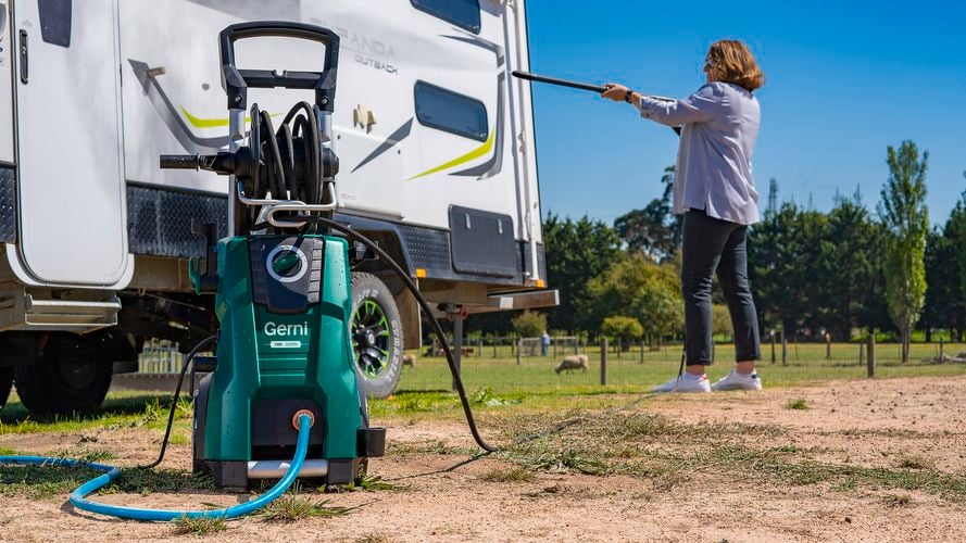 Person using a green Gerni pressure washer to clean the side of a white caravan in an outdoor setting with trees and grass in the background.
