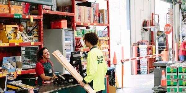 A Bunnings Trade specialist carrying lengths of timber talking to a Bunnings team member at a warehouse checkout