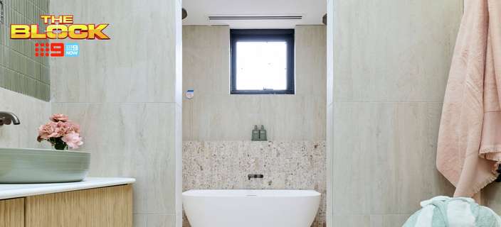 Modern neutral bathroom with a freestanding white bath, timber vanity, green basin, light tiled walls, folded towels and soft pink accents. “The Block” logo appears in the upper-left corner.