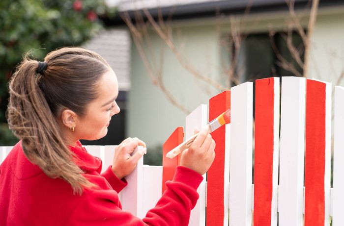 Painting alternating red and white stripes on a picket fence gate with a brush.