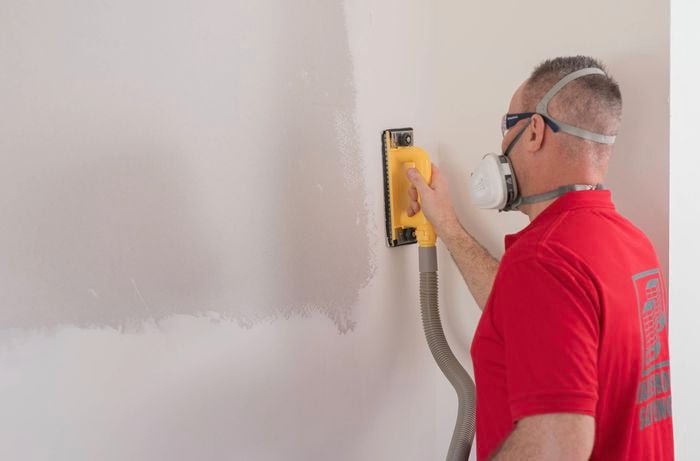 A Bunnings Team Member, wearing safety goggles and a dust mask, sands a wall to smooth out any bumps.